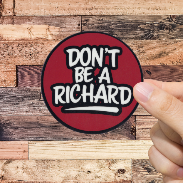 Hand holding a round sticker with 'Don't Be A Richard' text against a wooden background