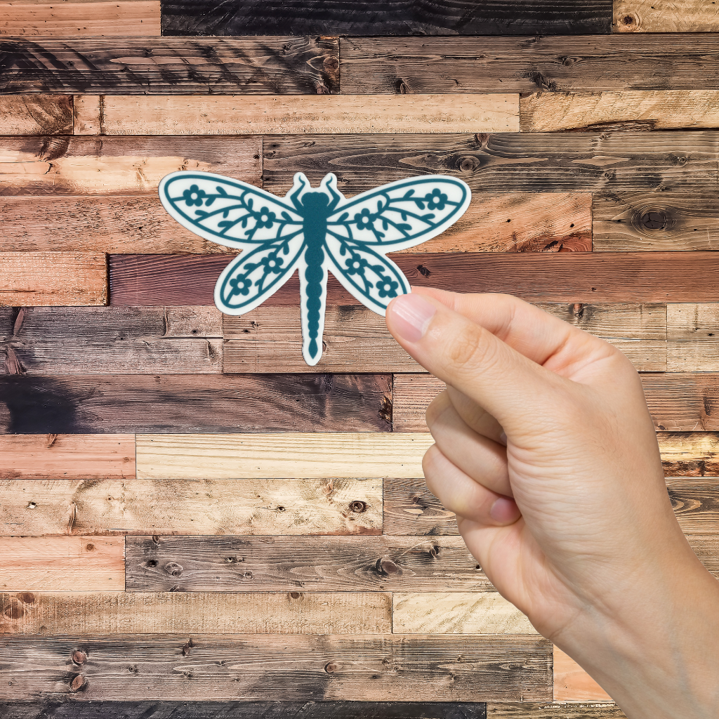 Hand holding a teal dragonfly sticker against a wooden background