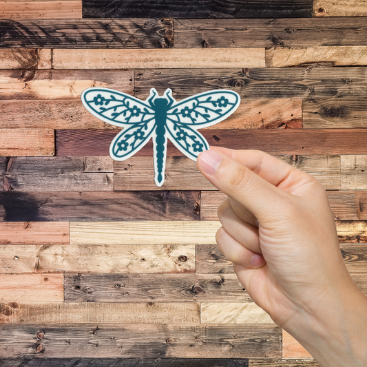 Hand holding a teal dragonfly sticker against a wooden background