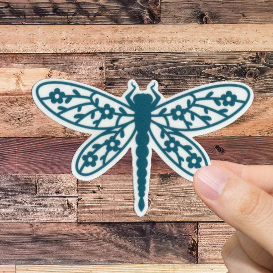 Hand holding a teal dragonfly sticker against a wooden background