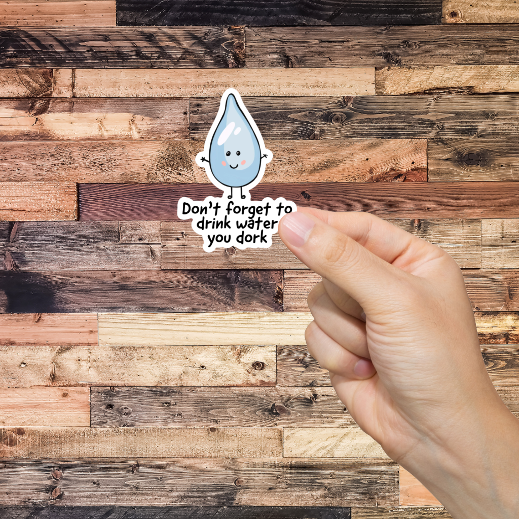 Hand holding a sticker with a water droplet and text 'Don't forget to drink water you dork,' against a wooden background