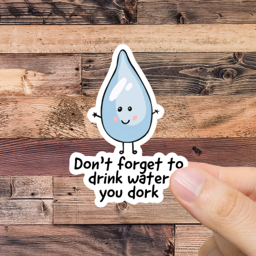 Hand holding a sticker with a water droplet and text 'Don't forget to drink water you dork,' against a wooden background