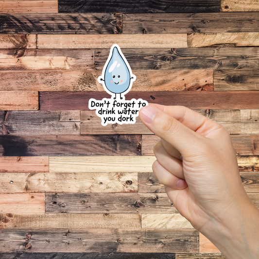 Hand holding a sticker with a water droplet and text 'Don't forget to drink water you dork,' against a wooden background