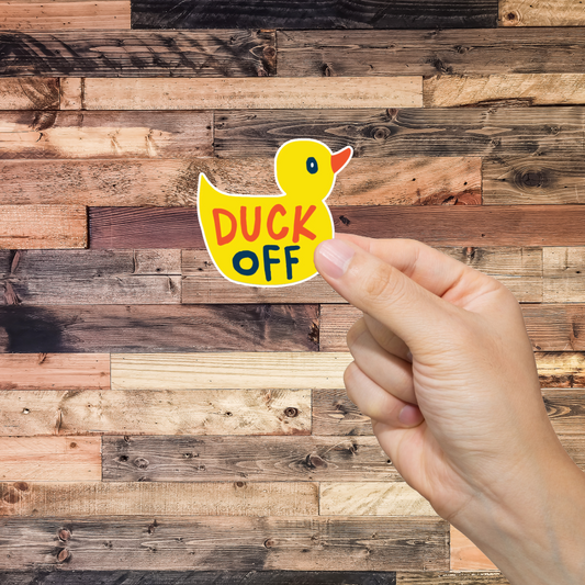 Hand holding a sticker with a duck and 'DUCK OFF' text against a wooden background