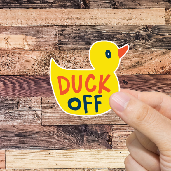 Hand holding a sticker with a duck and 'DUCK OFF' text against a wooden background