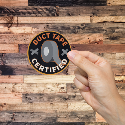 Hand holding a 'Duct Tape Certified' sticker against a wooden background