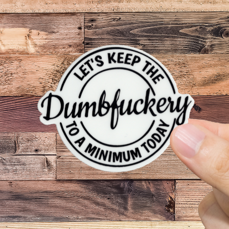 Hand holding a sticker with text 'let's keep the dumbfuckery to a minimum today.' against a wooden background