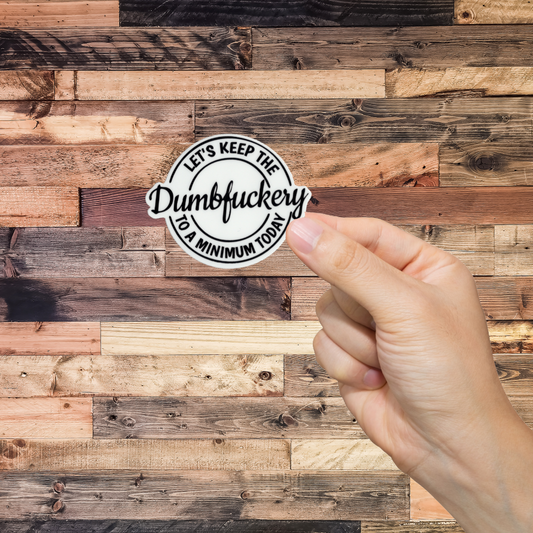 Hand holding a sticker with text 'let's keep the dumbfuckery to a minimum today.' against a wooden background
