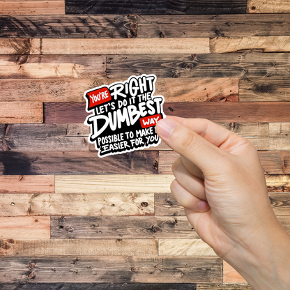 Hand holding a sticker with text 'You're Right Let's do it the Dumbest way possible' against a wooden background