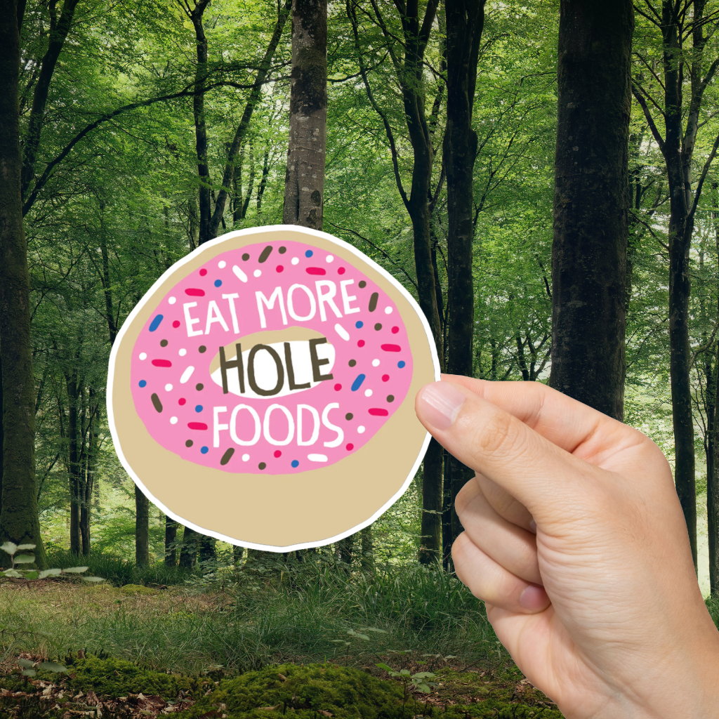 Hand holding a sticker with 'Eat More Hole Foods' text against a forest background