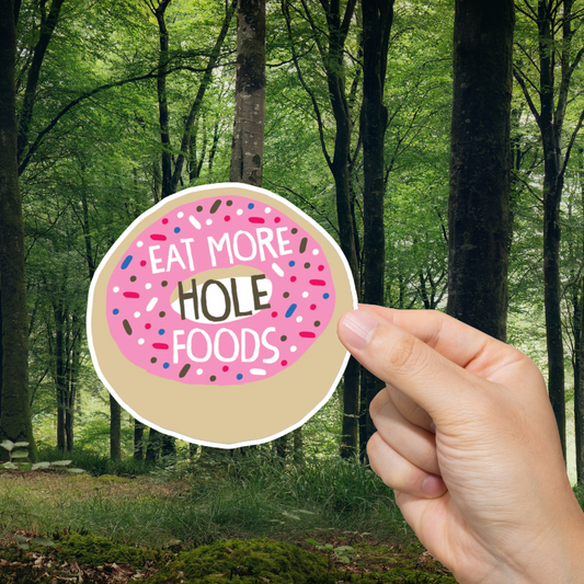 Hand holding a sticker with 'Eat More Hole Foods' text against a forest background