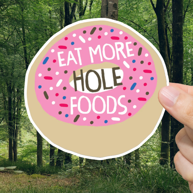 Hand holding a sticker with 'Eat More Hole Foods' text against a forest background