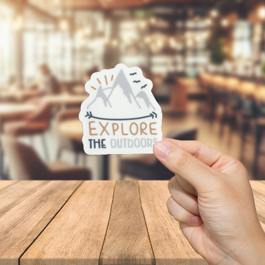 Hand holding a 'Explore The Outdoors' sticker over a blurred restaurant background