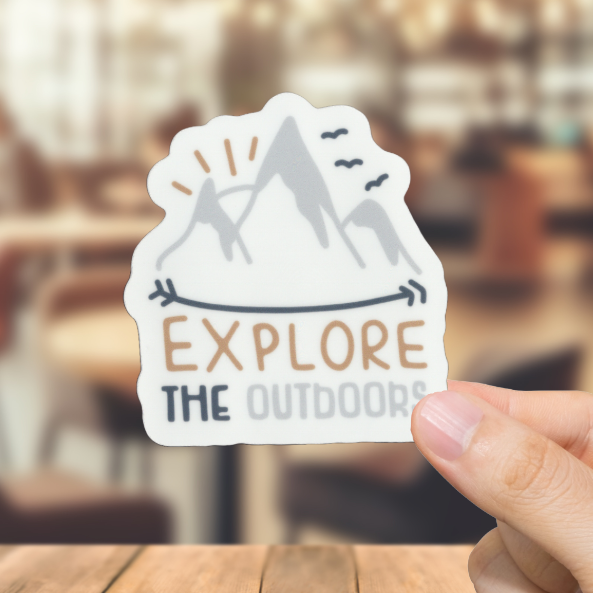Hand holding a 'Explore The Outdoors' sticker over a blurred restaurant background